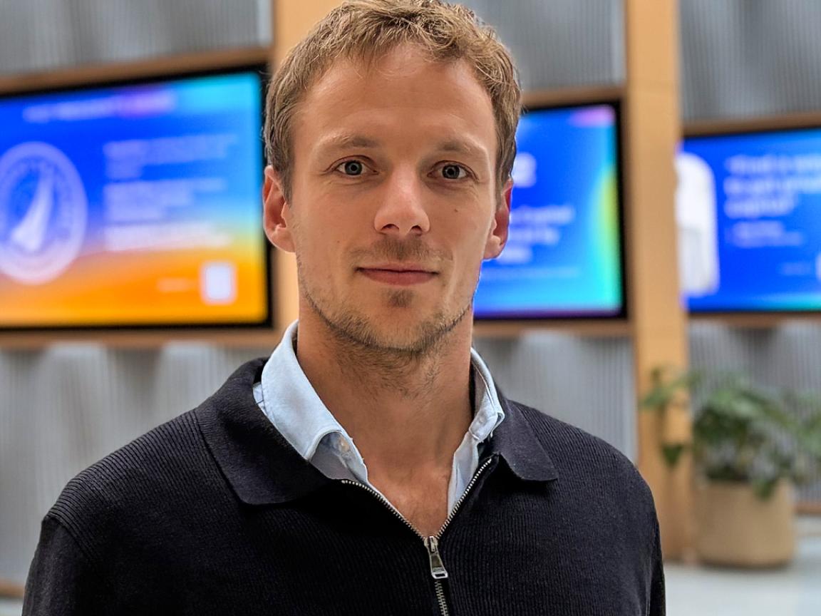 Jonas Wittern - Porträtfoto eines Mannes in einem Innenraum, der in die Kamera blickt, mit unscharfen Bildschirmen im Hintergrund.