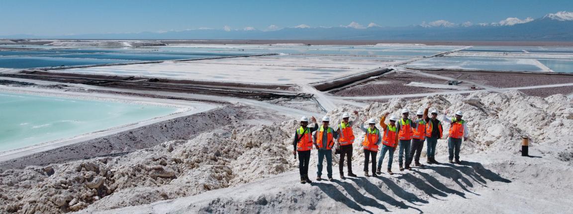Group of people with safety helmets and bright orange safety vests standing on a hill in a salt mining area with large evaporation ponds; in the background, mountains and a clear blue sky are visible.