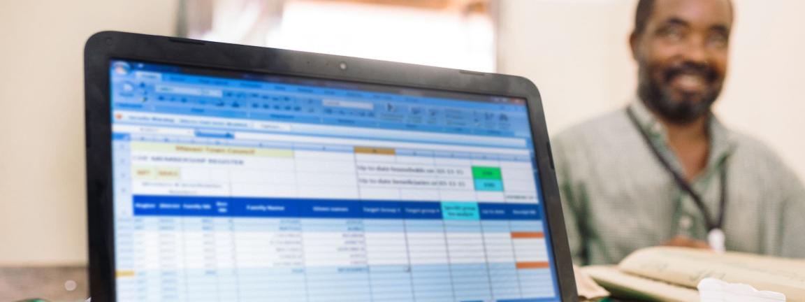 Auf dem Bildschirm eines Laptops ist ein Tabellenprogramm zu sehen. Der Laptop steht auf einem Tisch mit vielen Materialien; auf der anderen Seite sitzt ein freundlich lächelnder Mann.