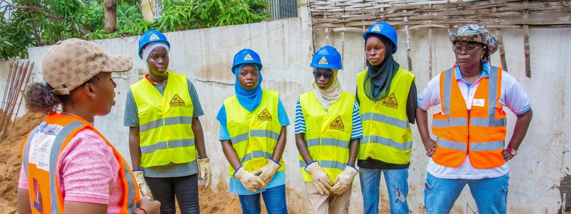 Eine Gruppe von fünf Frauen in neonfarbenen Warnwesten und Arbeitskleidung bekommen eine Einweisung von einer weiteren Frau im Vordergrund. Die Personen stehen auf einem sandigen Untergrund auf einer Baustelle.
