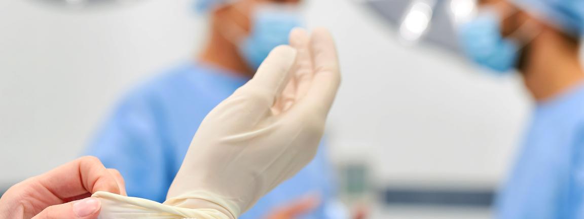 A medical professional is putting on sterile gloves in an operating room, with two other people in surgical attire blurred in the background.