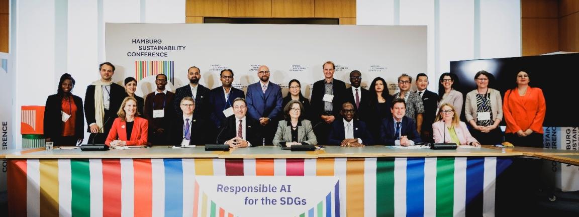 Eine Gruppe von Menschen steht in zwei Reihen vor einem langen Tisch, an dem ein bunt gestreifter Banner befestigt ist. 