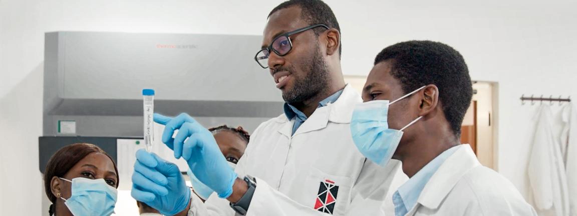 A man in a lab coat is showing a group of colleagues how to handle a test tube with a sample, emphasizing teamwork and knowledge sharing in the laboratory.