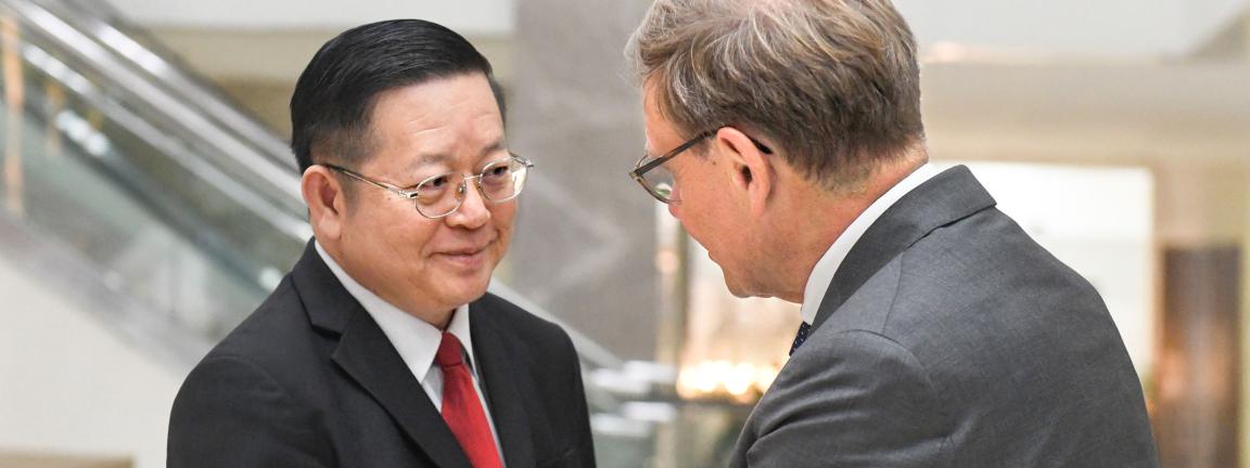 Zwei Männer in Anzügen begrüßen sich in einem modernen Gebäude mit Rolltreppe im Hintergrund bei einem formellen Treffen.