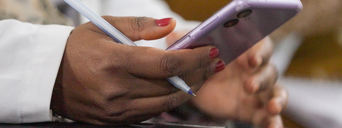 Close-up of a hand holding a purple smartphone while the other hand holds a pen. A notebook is visible in the background, with the focus on the hands.