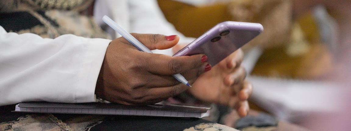 Nahaufnahme einer Hand, die ein Smartphone in lila Farbe hält, während die andere Hand einen Stift hält. Im Hintergrund ist ein Notizbuch sichtbar, und der Fokus liegt auf den Händen.