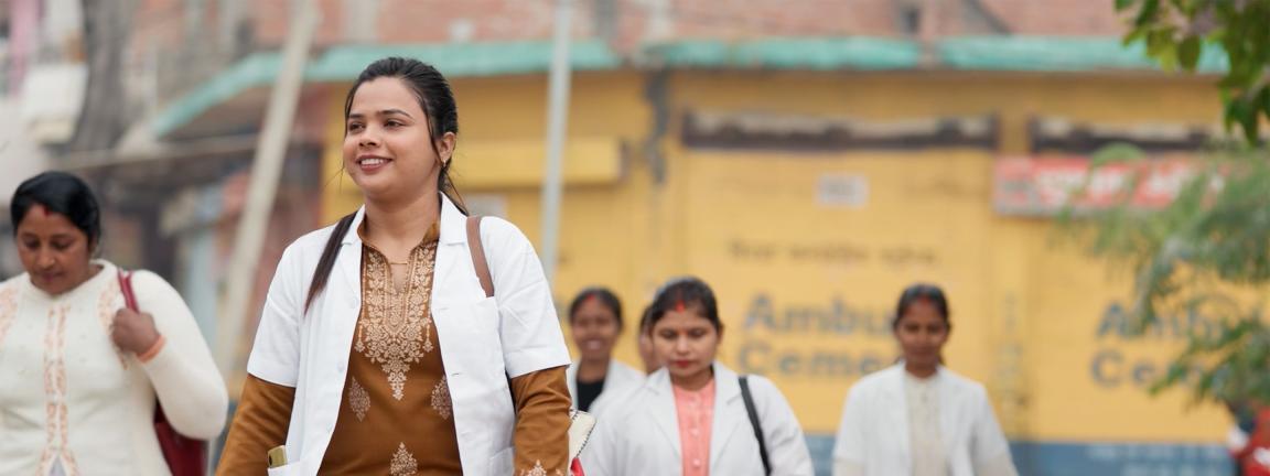 Eine Gruppe von Frauen in weißen Kitteln geht gemeinsam über eine Straße, angeführt von einer Frau in brauner Kleidung, vor einem unscharfen Gebäude im Hintergrund.