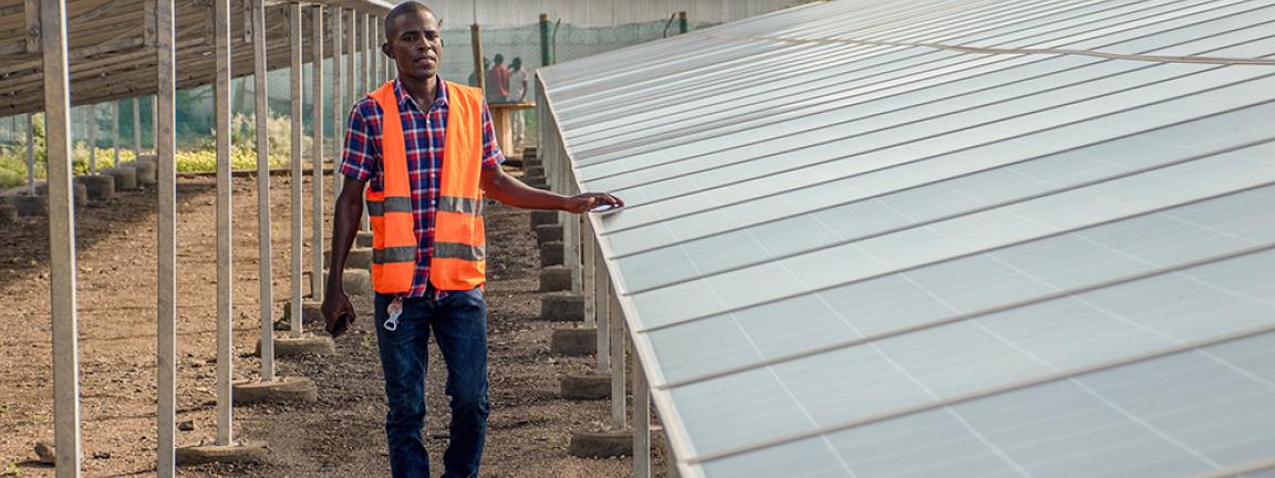 Mann mit orangefarbener Warnweste und kariertem Hemd steht neben einer Reihe von Solarpaneelen auf einem Freigelände mit Metallkonstruktionen und Gebäuden im Hintergrund.