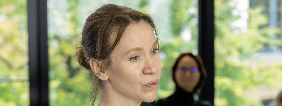 Frau mit zusammengebundenen Haaren hält ein Dokument in der Hand und spricht in einem hellen Seminarraum mit großen Fenstern, während im Hintergrund weitere Personen sitzen.