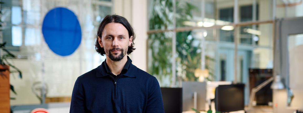 Ein Mann in einem dunkelblauen Pullover mit Reißverschluss sitzt mit gefalteten Händen in einem modernen, begrünten Büro und blickt ruhig in die Kamera.