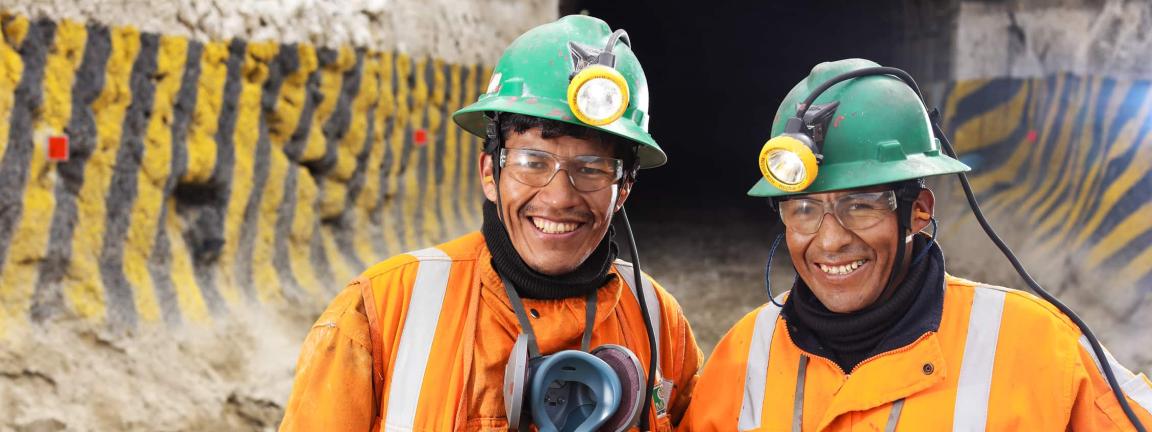 Zwei lächelnde Bergarbeiter in orangefarbener Schutzkleidung mit grünen Helmen und Stirnlampen stehen vor einem Stolleneingang.