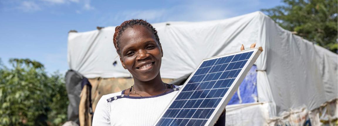 A woman smiles at the camera while holding a solar panel in front of a makeshift shelter.