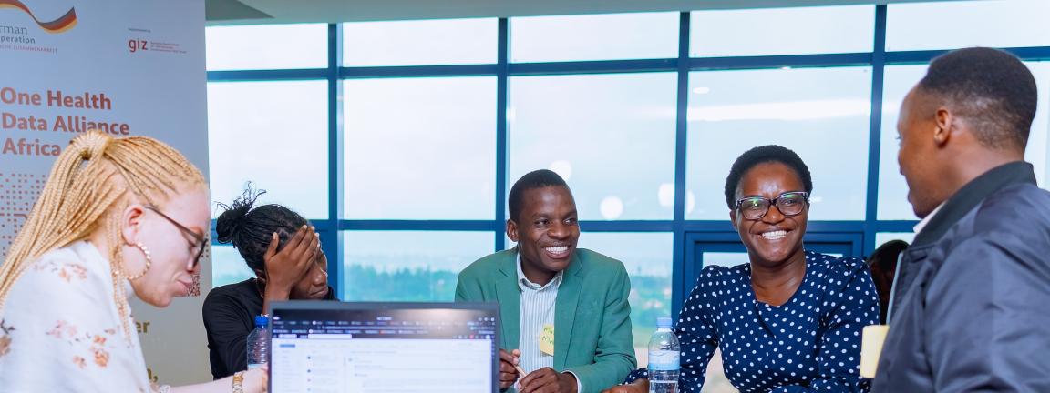 Several people sit around a table in a bright room with a laptop and documents, talking, with a “One Health Data Alliance Africa” banner in the background.