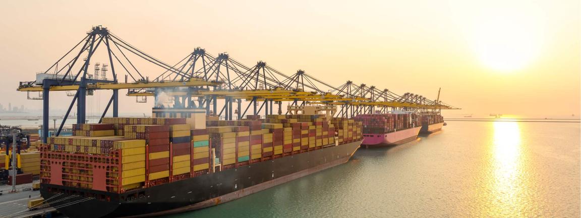 Ein Containerschiff liegt bei Sonnenuntergang an einem Terminal und wird von mehreren Hafenkränen beladen, während Container in Reihen gestapelt sind.
