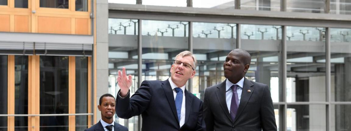Three men in suits stand in a modern building while the man in the middle gestures upward with a raised hand.