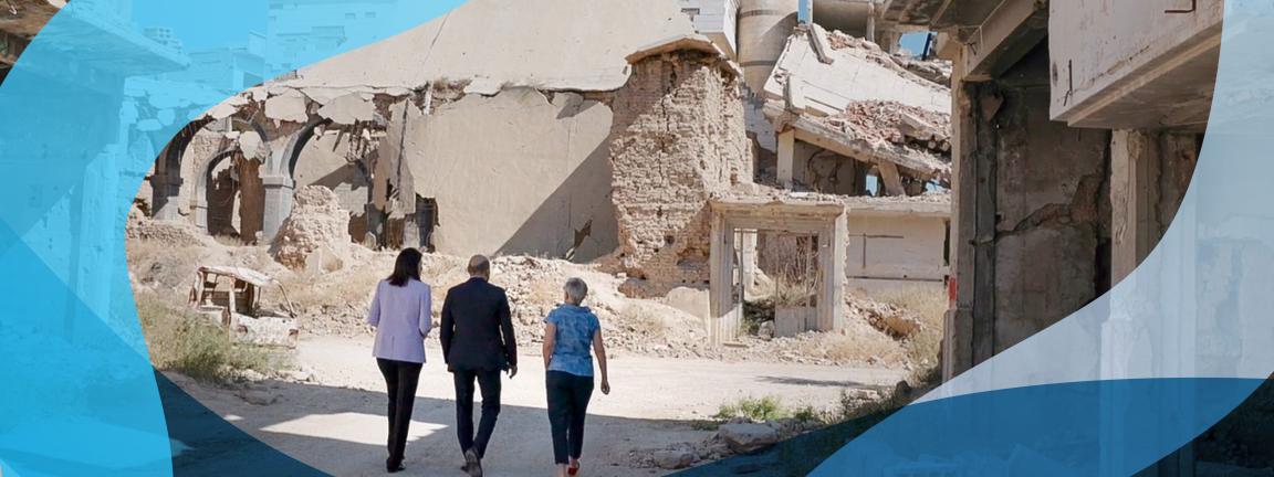 Three people walk along a street. All of the buildings are severely damaged. 