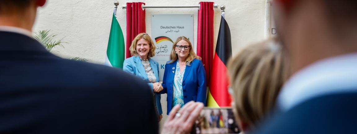 Zwei Frauen schütteln sich bei einer Veranstaltung vor einer Tafel mit der Aufschrift „Deutsches Haus“ die Hand, flankiert von roten Vorhängen und zwei Fahnen, während unscharfe Personen im Vordergrund zusehen und jemand ein Foto mit dem Smartphone macht.