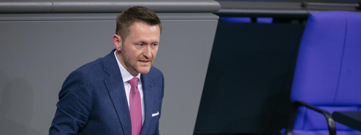 Ein Mann im blauen Anzug mit pinker Krawatte spricht gestikulierend an einem Rednerpult in einem Plenarsaal.