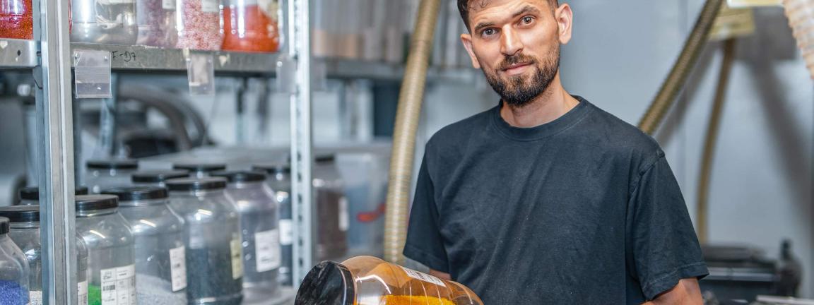Ein bärtiger Mann in dunklem T-Shirt und schwarzen Arbeitshandschuhen hält ein Glas mit orangefarbenen Kunststoffgranulaten und blickt lächelnd in die Kamera, im Hintergrund stehen Regale mit weiteren Behältern voller farbiger Granulate.