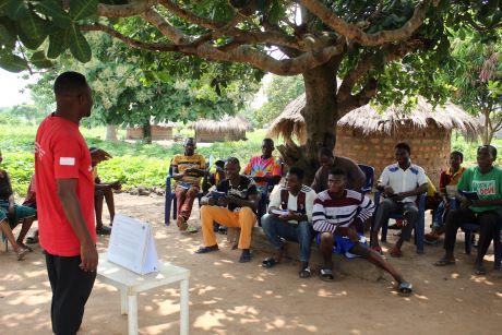 Ein Mann hält draußen einen Vortrag. Um einen Baum herum sitzen die Zuhörer.