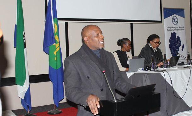 Man giving a speech, 2 women behind him sitting at a table.