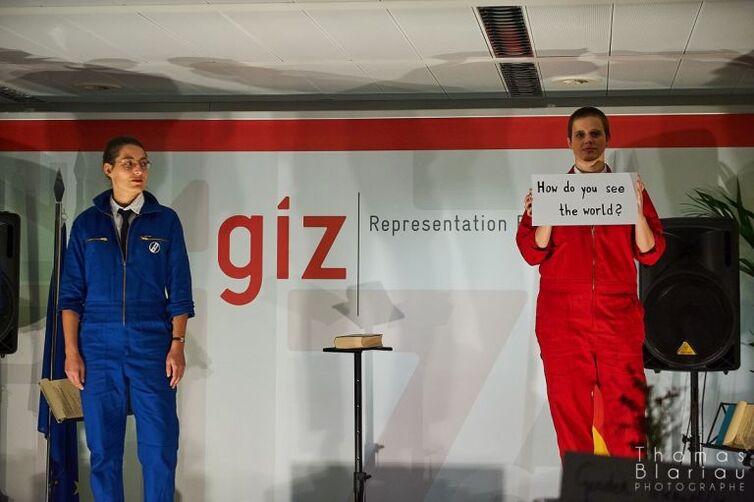 Links steht eine Person in einem blauen Overall, rechts steht eine Person in einem roten Overall. Diese hält ein Schild mit der Beschriftung "How do you see the world" hoch