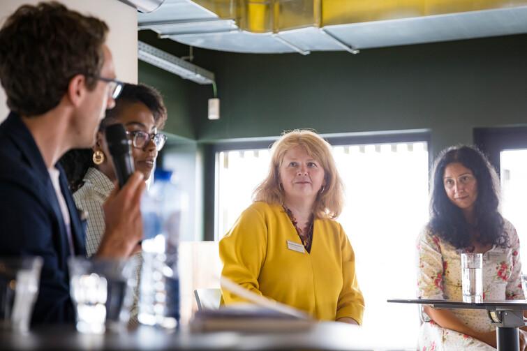 Frau in einem gelben Oberteil schaut nach links zu den beiden Moderatoren