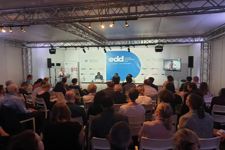 Kleine Bühne auf der eine Person an einem Tisch sitzt. Davor sitzen viele Menschen auf Stühlen im Raum verteilt