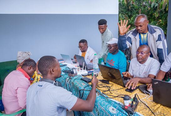 Vier Personen sitzen mit jeweils einen Laptop am Tisch. Gegenüber und dahinter stehen noch mehr Personen.
