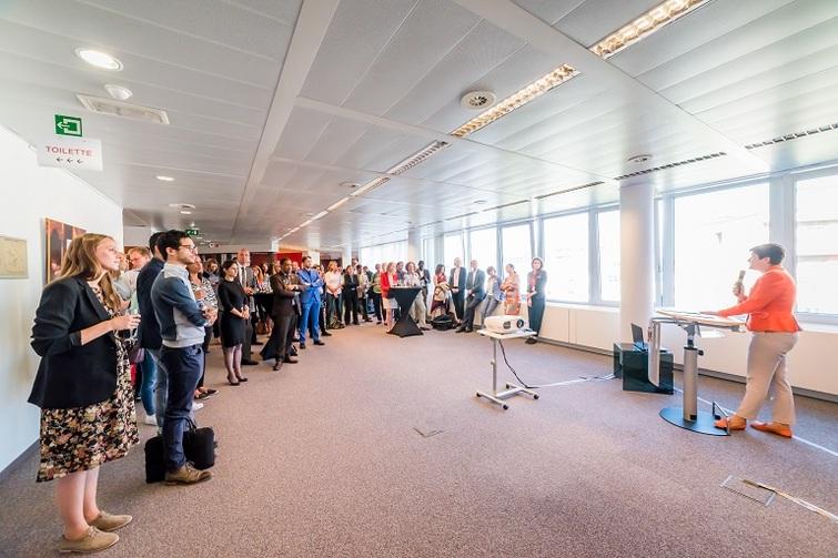 Viele Menschen stehen verteilt in einem Raum. Eine Person steht vorne an einem Tisch und hält ein Mikrofon