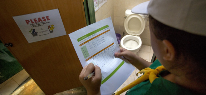 Eine Person in Uniform steht vor einer geöffneten Toilette und bearbeitet handschriftlich eine Check-Liste.