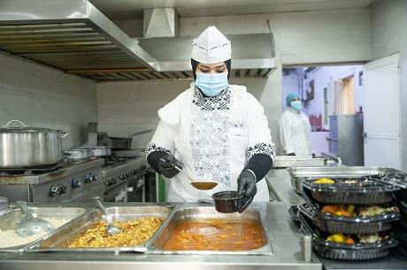 A woman in professional chef attire serving food in a commercial kitchen.