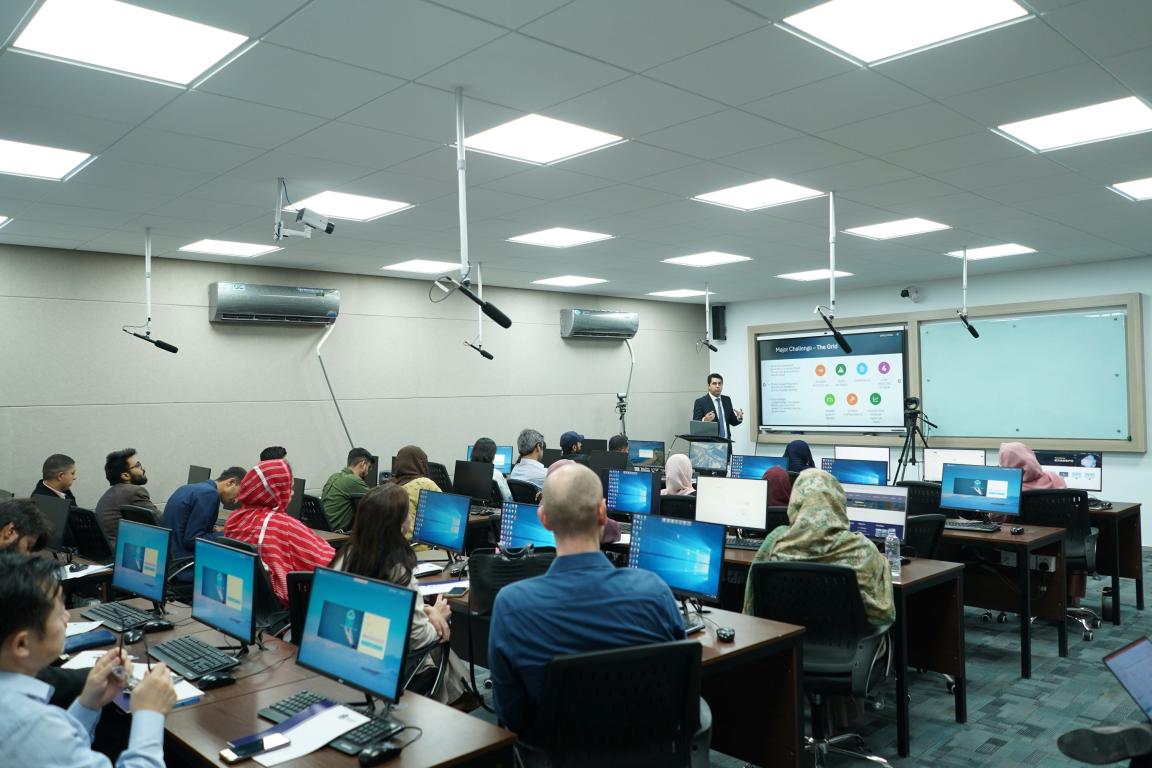 A trainer leads a hands-on technical session in a computer lab, where many participants are seated at individual desks and monitors.