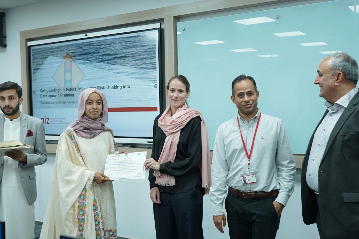A female participant receives a training certificate from PGCEP members in front of a screen displaying a risk planning slide.