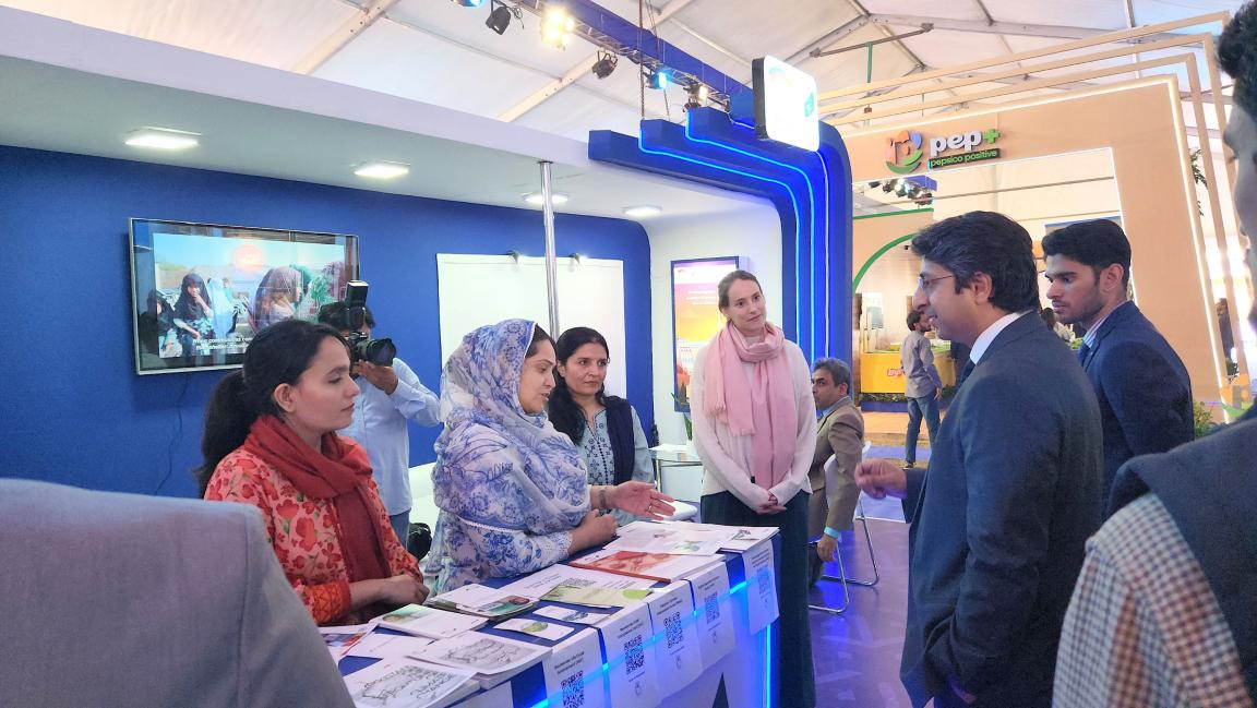 A group, including the PM's Coordinator, interacts with PGCEP staff at an exhibition booth featuring a display unit and brochures.