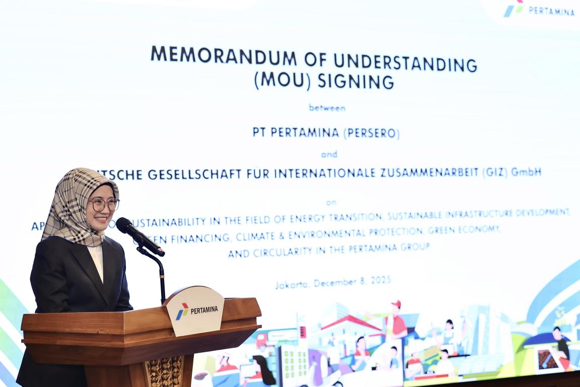 Woman with White/Black Veil and glasses smiling speaking at the Podium with the Backdrop of Pertamina x GIZ MoU Signing