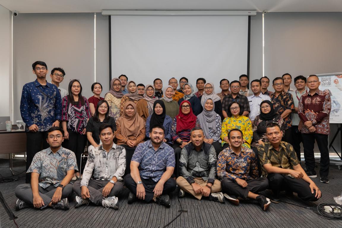 Group photograph of approximately 35 people posing indoors in front of a blank projection screen and whiteboard. Participants wear a mix of casual and traditional attire, with some seated on the floor and others standing behind.