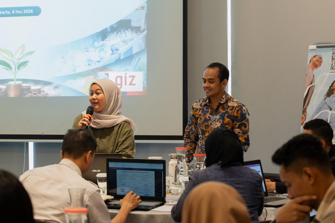 Photograph of a business meeting or workshop showing a woman speaking into a microphone and a man standing beside her in front of a presentation screen. Attendees sit around a table with laptops and documents, while the screen displays a slide with text and logos, indicating a professional or educational setting.