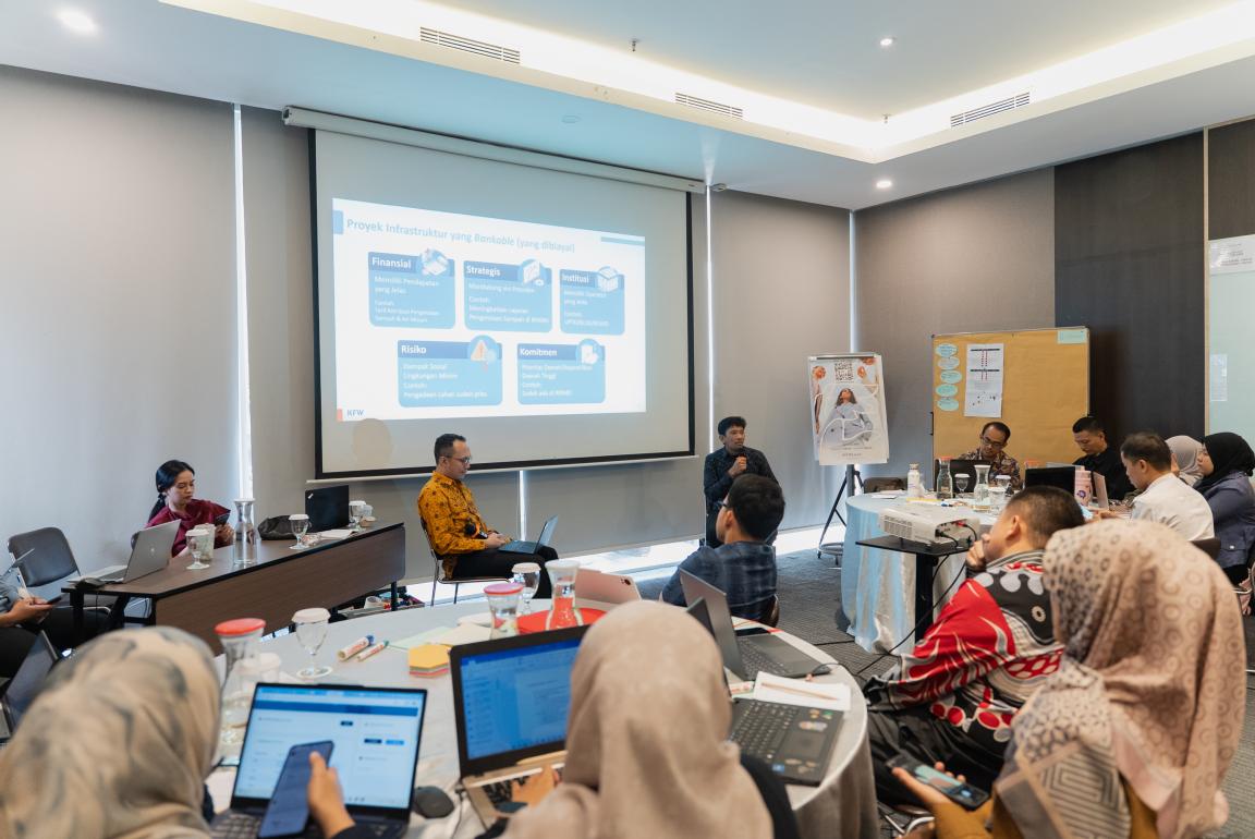 Photograph of a business meeting or workshop with participants seated around a conference table, some using laptops, and a presenter speaking in front of a projected slide. The slide displays a flowchart with labeled boxes in blue and white, outlining process information related to financial, strategies, institution, commitment, and risk categories.