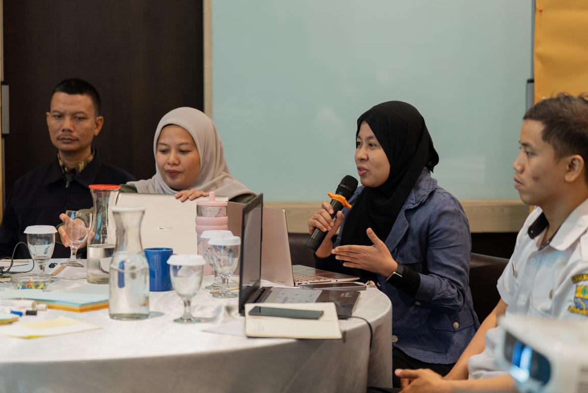 Photograph of a group meeting around a round table with laptops, notebooks, and water glasses, where one person wearing a hijab and denim jacket is speaking into a microphone.