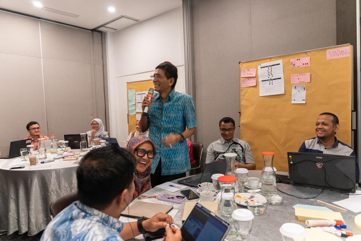 Photograph of a business meeting or workshop with participants seated around tables, some using laptops and others listening to a standing speaker holding a microphone. A large brown board with pink sticky notes and printed charts is visible in background, indicating brainstorming or project planning activities.