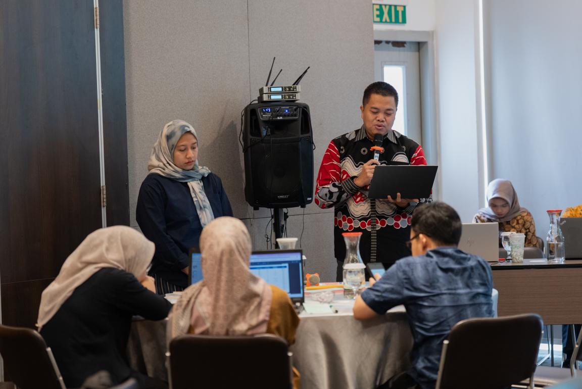 Photograph of a group meeting or workshop in a conference room with one person standing and speaking into a microphone while holding a laptop, and others seated around a table with laptops and notebooks.