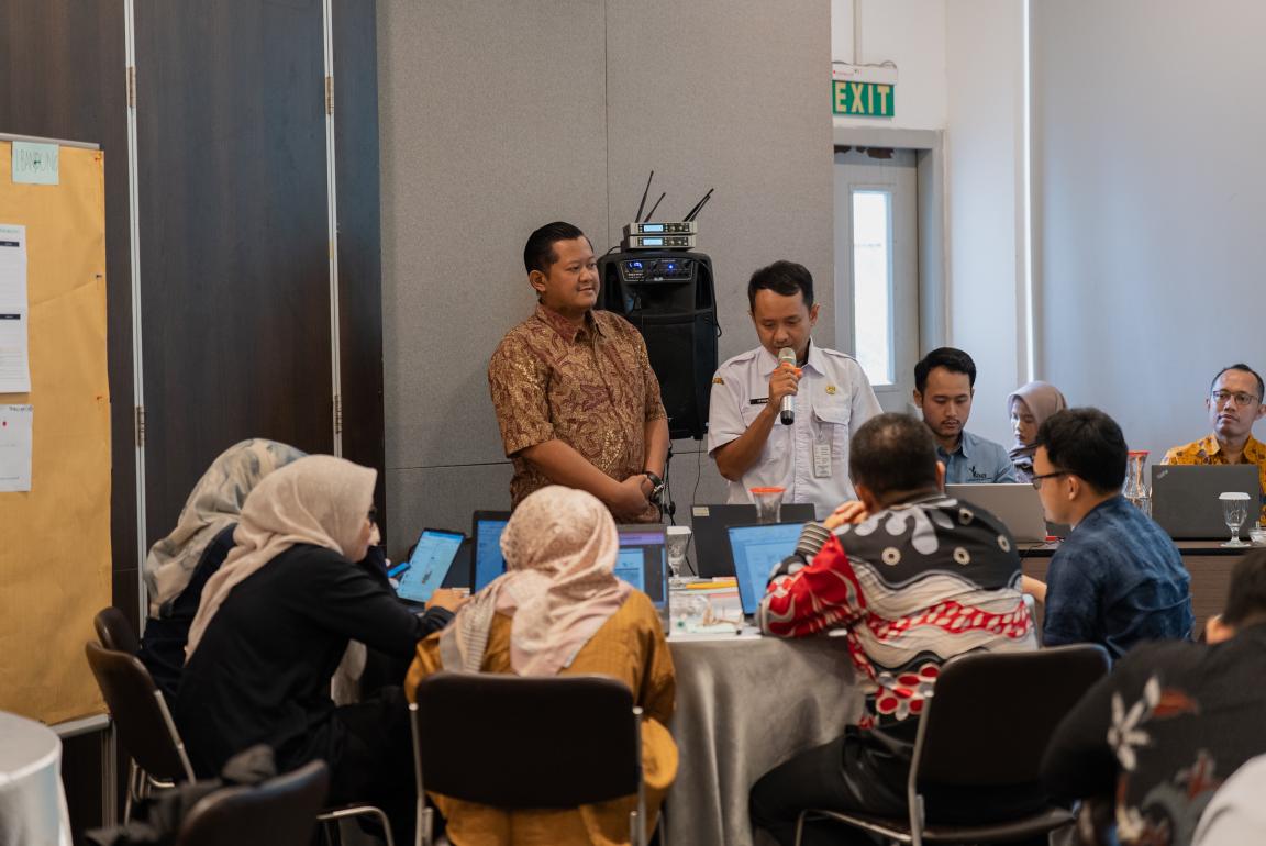 Photograph of a group meeting in a conference room where two men stand near a speaker system, one holding a microphone, while others sit around a table with laptops and notebooks.