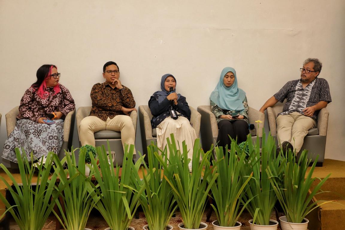 Five people sitting in a row on chairs against a plain wall, with several potted plants arranged in front of them. The individuals wear casual and traditional clothing, with some women wearing headscarves.