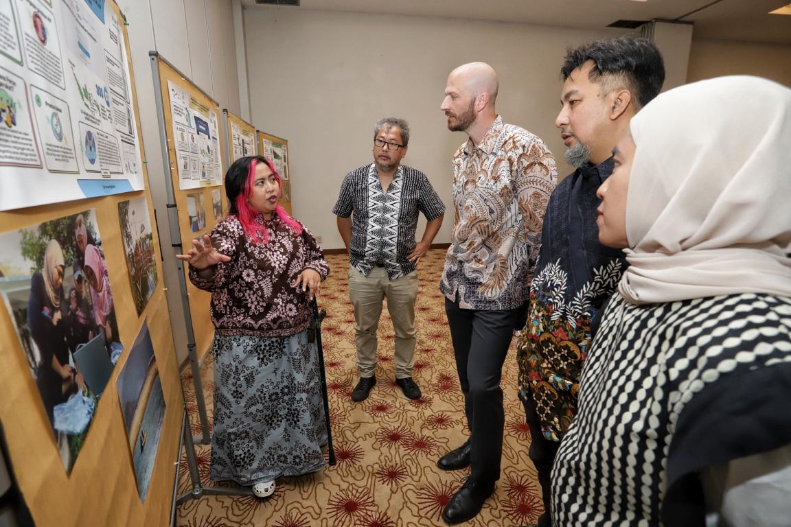 A group of five people standing and observing a series of informational posters displayed on a wall in a carpeted room. One woman appears to be explaining content on the posters, which include text, images, and diagrams, while others listen attentively.