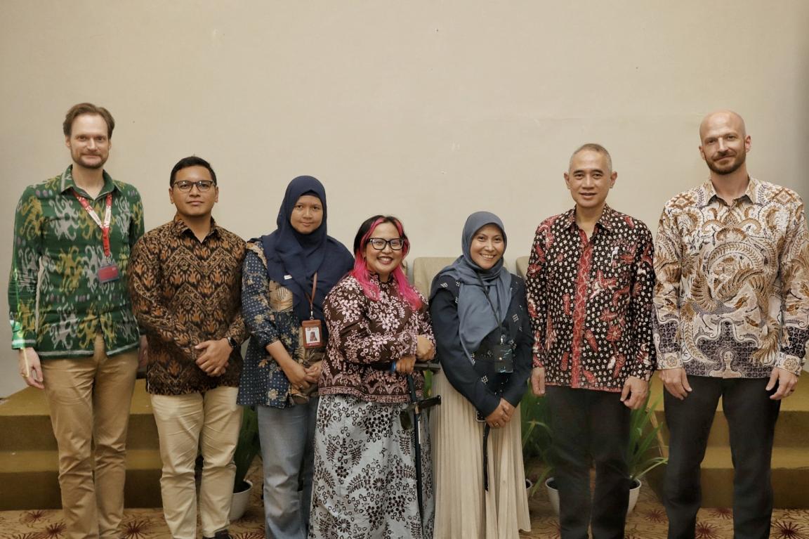 Photograph of seven people, three women and four men, standing indoors and wearing traditional patterned clothing in various colors, including green, brown, blue, and white.