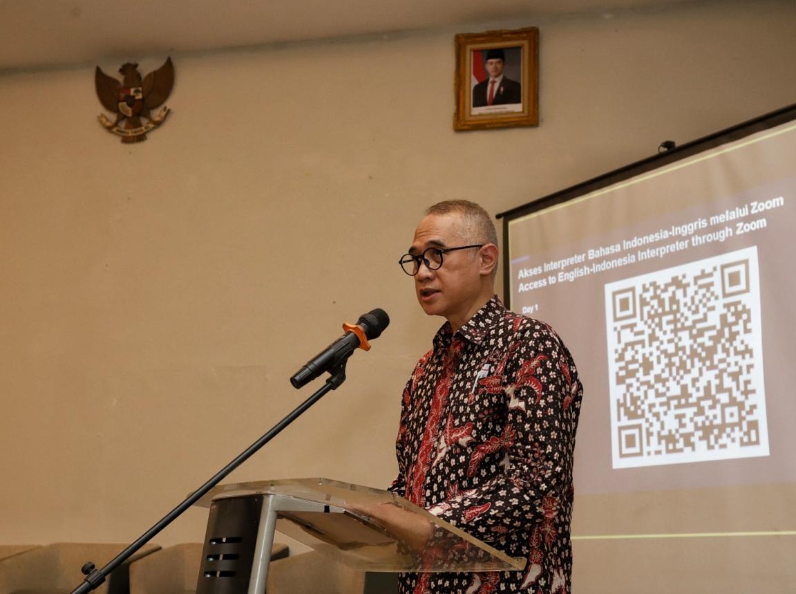 A man speaking at a podium with a microphone in front of a projection screen displaying a QR code and bilingual text about accessing English-Indonesian interpretation through Zoom.