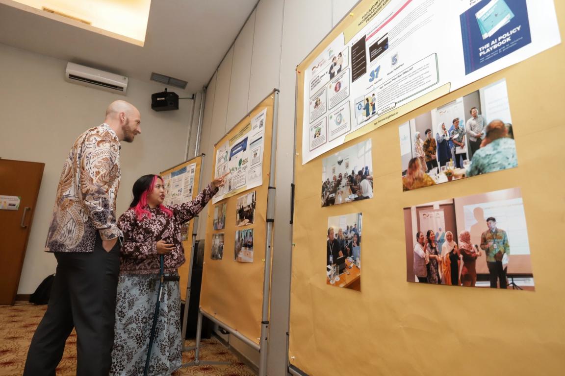 Two people examining a display board featuring multiple posters and photographs of an exhibition. Display includes informational posters with text and images highlighting project achievements, with the individuals engaged in discussion about the content.