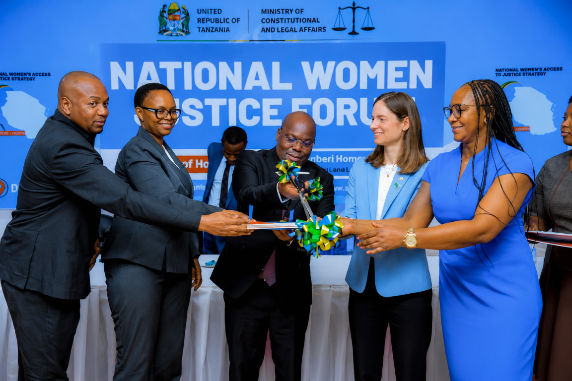A group of officials and guests participate in a ceremonial ribbon-cutting during the launch of the National Women Justice Forum in Tanzania. The event takes place on a stage with a large backdrop displaying the forum's name alongside the logos of the United Republic of Tanzania and the Ministry of Constitutional and Legal Affairs. Six people stand at the front, smiling as they jointly cut a green, yellow, and blue ribbon tied around a publication or plaque.