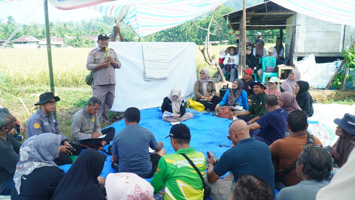 Photograph of an outdoor community meeting held under a striped canopy in a rural setting with rice fields and a wooden structure in the background. Participants, including uniformed officers and civilians, sit on a blue tarp while one officer stands and speaks, indicating a discussion or briefing session.